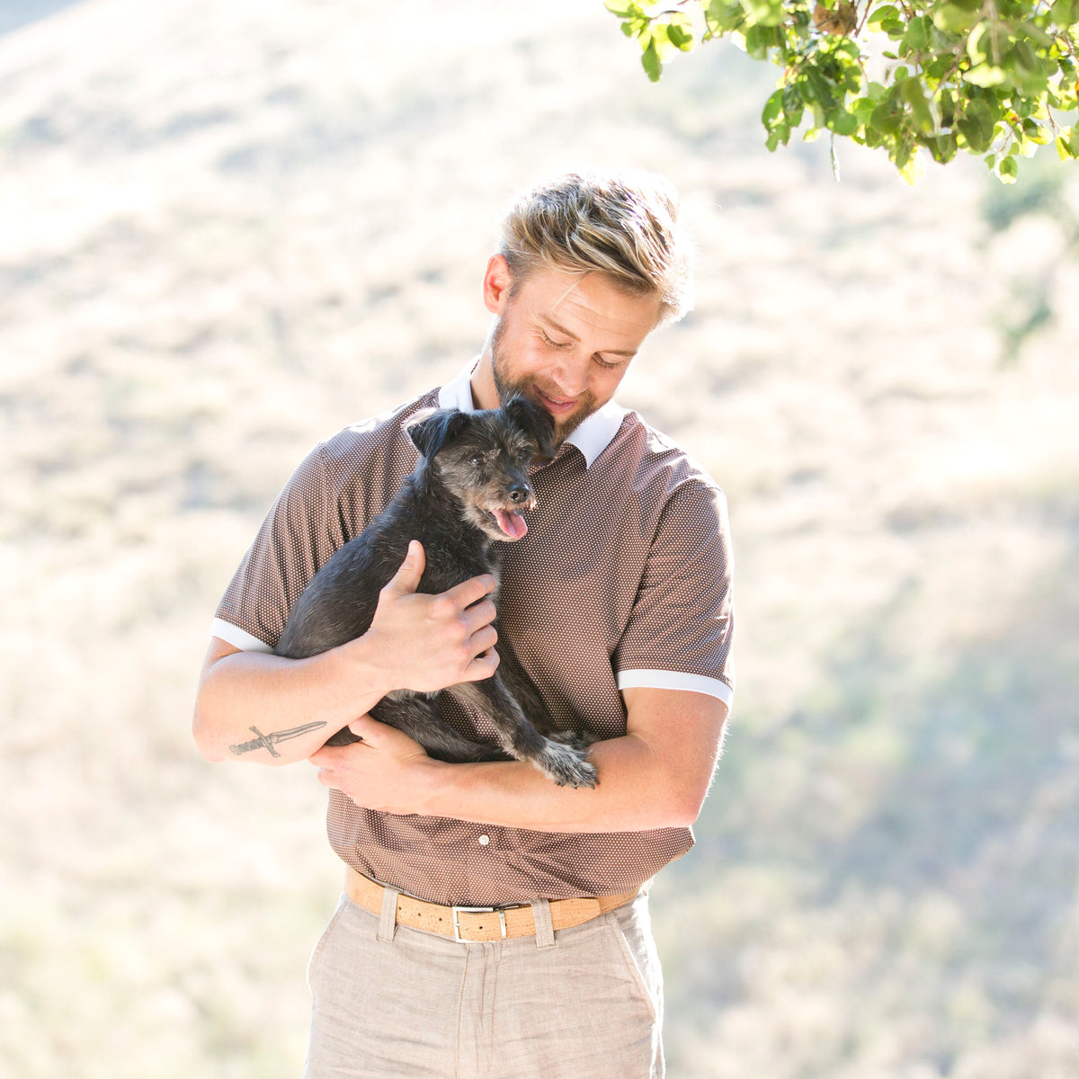 A man, dressed in a tucked brown patterned polo shirts, khaki shorts and cork belt stands outdoors, lovingly holding a little black dog. 