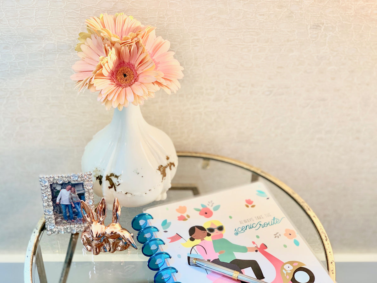 Atop a gold and glass table sits a milk glass vase with peach daisies, two rose gold rabbits, a small framed photo of a couple and a daily journal. 