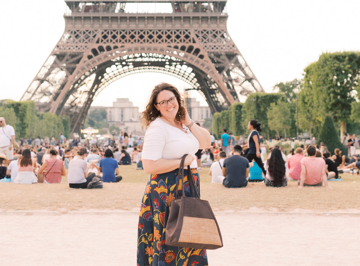 A lovely brunette woman cheerfully stands in front of Paris' Eiffel Tower carrying a chocolate brown and snake print cork tote handbag. 