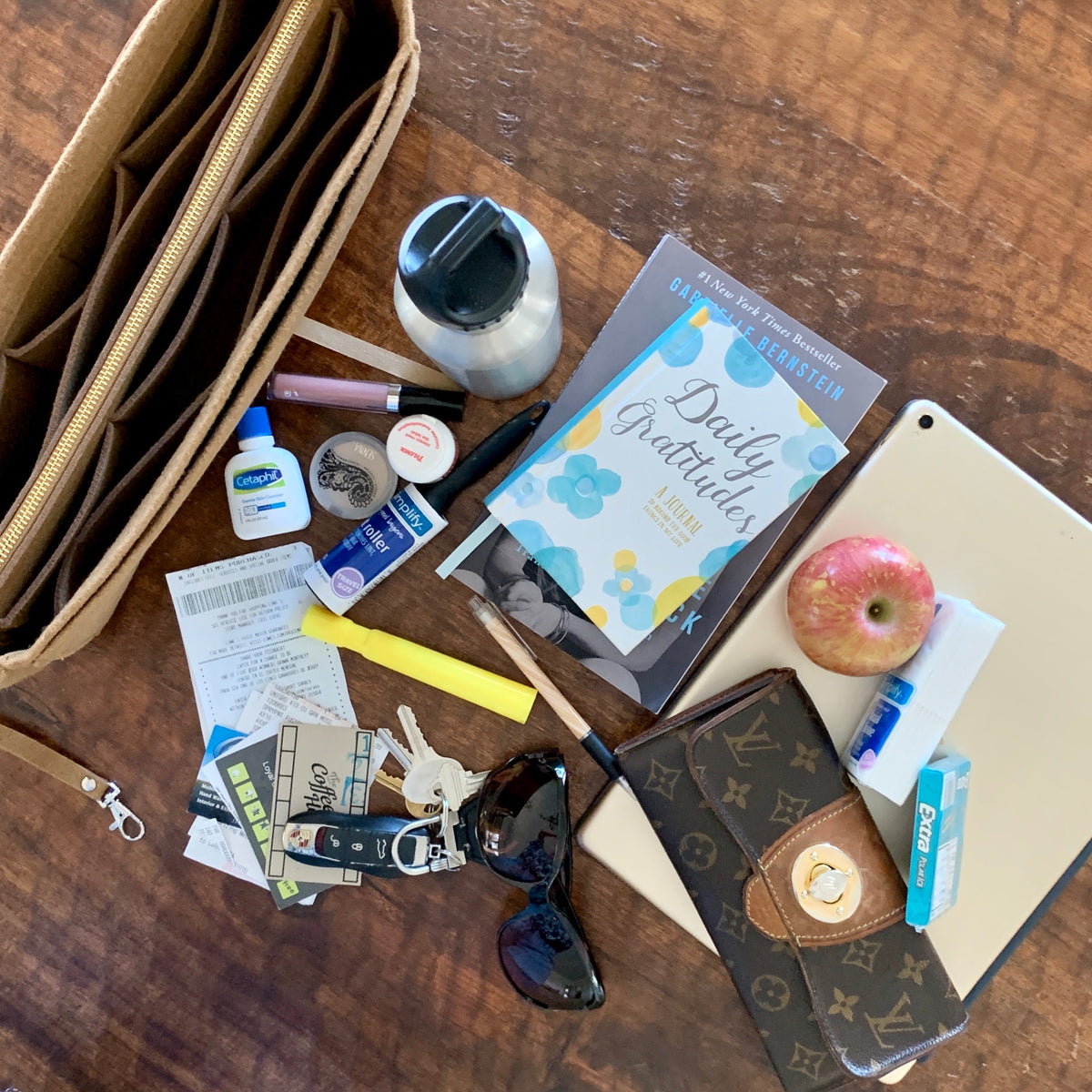 A purse organizer sits on a wooden table along with a wallet, iPad, apple, tissue, gum, pen, keys, lotion, aspirin, money, sunglasses and a journal at the ready to be organized. 
