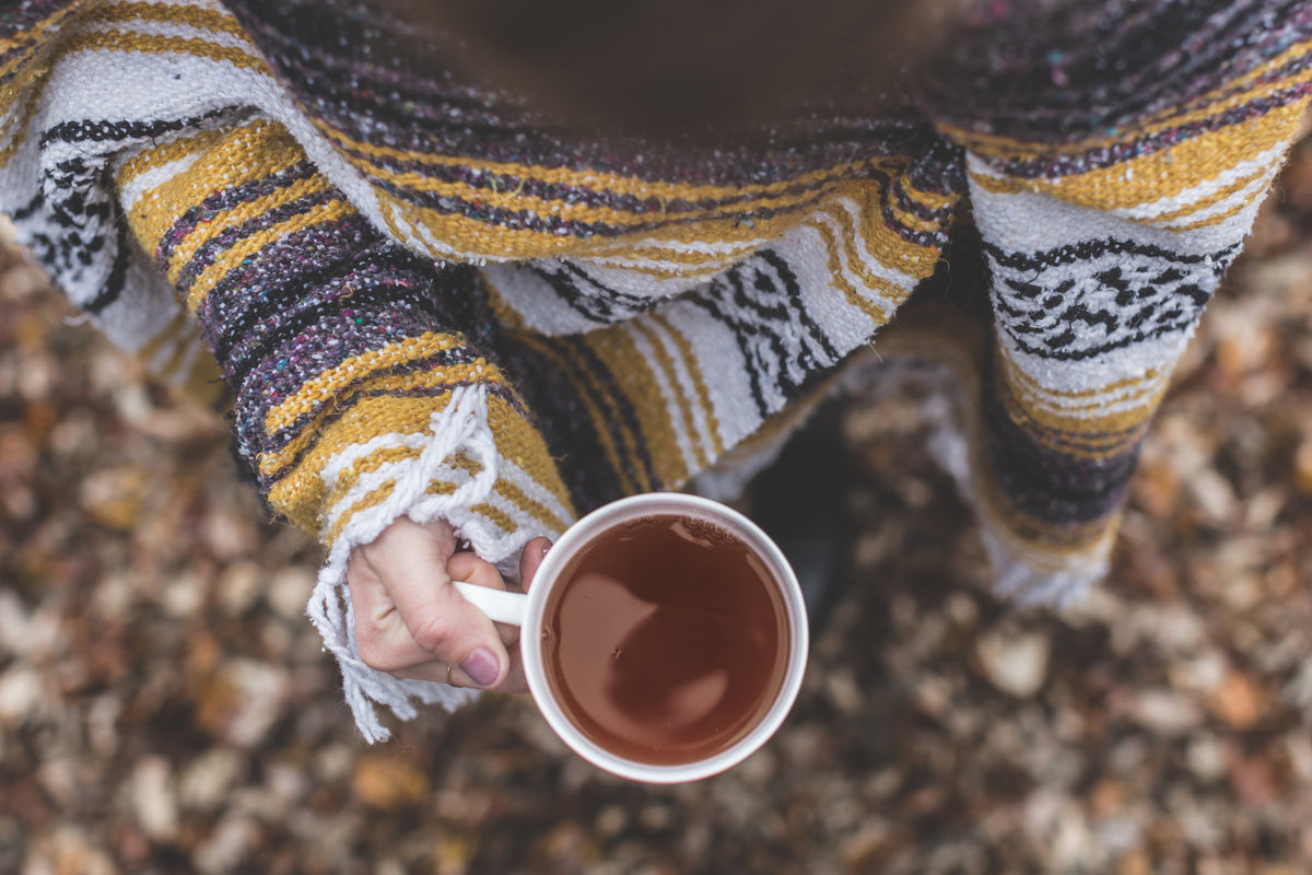A woman stands on fall leaf filled ground wrapped in a yellow, grey, black and white wool blanket with a cup of warm tea in her hand. 