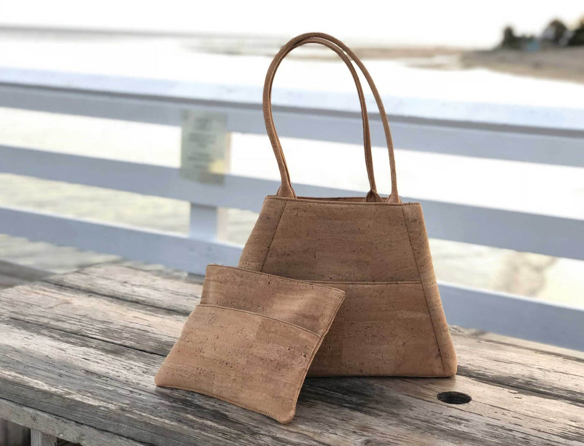 Two unique cork bags, a wristlet and shoulder bag or tote purse, made in the of cork fabric sit on an old wood picnic table on a beach pier. 