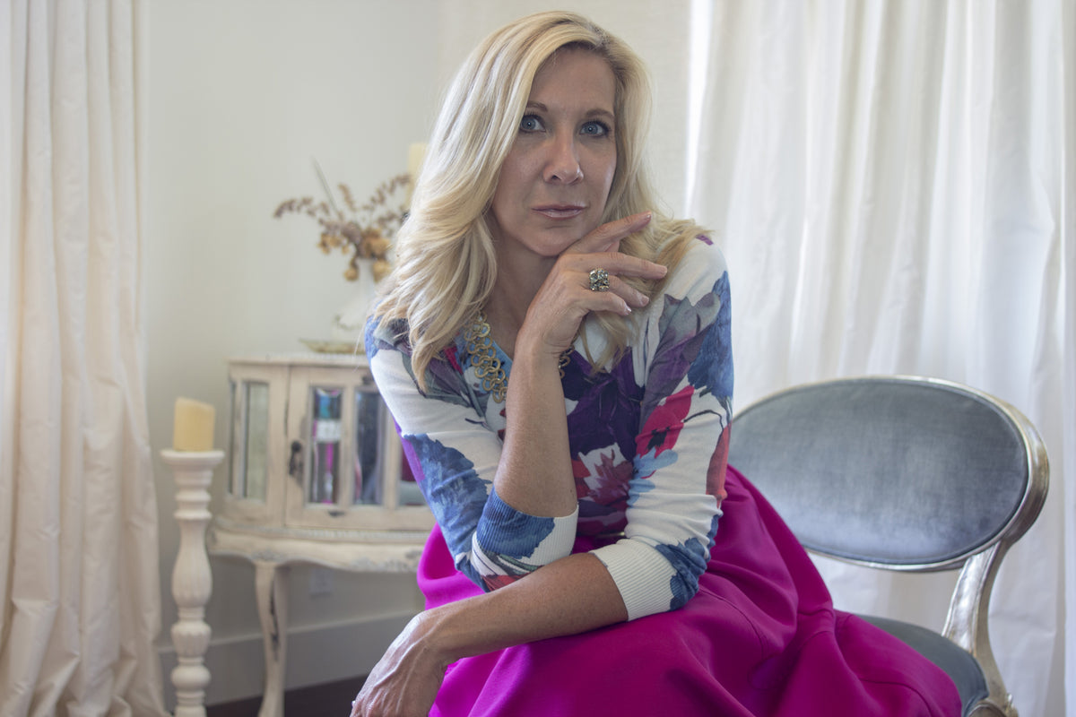 Personal stylist from Solvang, CA sits in room with cream walls and cream satin curtains wearing a beautiful brightly colored floral sweater and hot pink full skirt in a aqua satin and gold chair.  