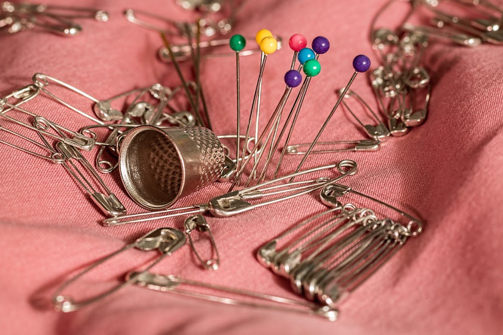 A silver thimble, safety pins and sewing pins sit atop a pink colored fabric 